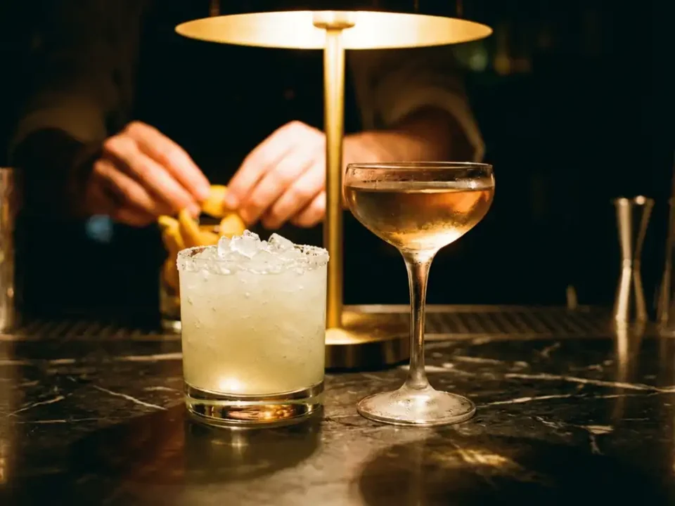 Gefrorene Margarita mit Salzrand und klassischer Martini auf Marmorbar, Barkeeper bereitet Garnituren im Hintergrund vor